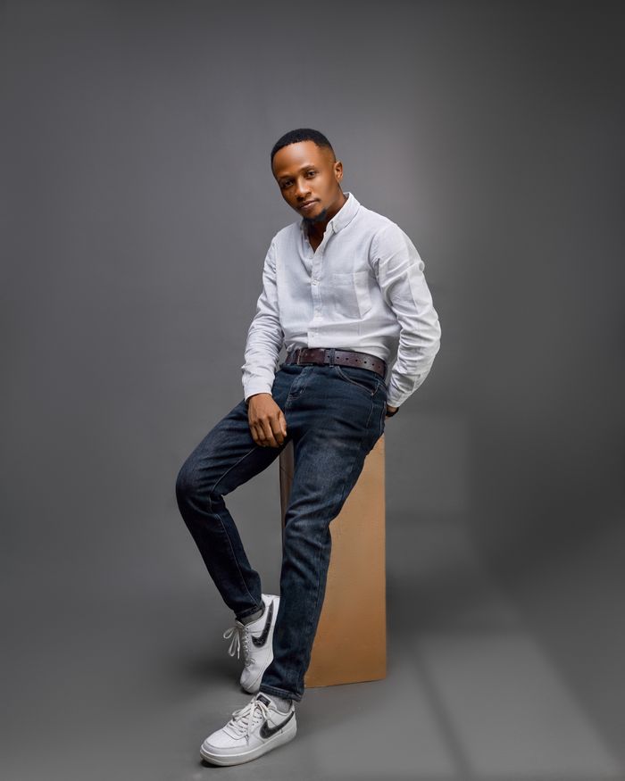 Fashionable male model posing in a studio wearing a white shirt, jeans, and sneakers against a gray background.